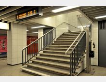 Eine U-Bahn-Station mit gefliesten Wänden, einer Treppe mit Metallgeländern, einem Deckenbildschirm mit Abfahrtszeiten und einem öffentlichen Münztelefon an der rechten Seite. Im Hintergrund sind rote Türen und ein Werbeplakat sichtbar.