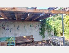 Holzüberdachter Außenbereich mit einer verwitterten Betonwand, die teilweise mit Farbspritzern bedeckt ist, und Kletterpflanzen auf der rechten Seite. Darunter stehen zwei Holzbänke, umgeben von Laub und Pflanzen.