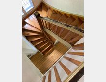 Holztreppe in einem engen Innenraum, die sich in einer kurvigen Form nach unten schlängelt. Die Stufen und das Geländer zeigen Gebrauchsspuren, und ein Fenster lässt Tageslicht auf die Holzoberflächen fallen.