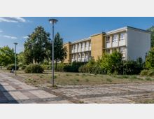 Ehemaliges Schulgebäude mit gelber und weißer Fassade, umgeben von Büschen, Bäumen und einer ungepflegten gepflasterten Fläche mit vereinzelten Laternen. Der Himmel ist klar und blau mit wenigen Wolken.