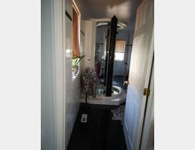 Ein Badezimmer mit schwarzen glänzenden Bodenfliesen, einer runden, modernen Duschkabine in der Ecke und einem Fenster mit Holzjalousien. Auf einem Handtuchhalter hängt Stoff mit Zebramuster, und ein kleiner Pflanzenkübel steht auf der Fensterbank.