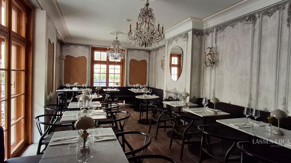 Eleganter Restaurant-Speisesaal mit hohen Holzfenstern, marmorierten grauen Wandpaneelen und dunkler Sitzbank entlang der Wand. Mehrere gedeckte Tische mit schwarzen Stühlen stehen auf Fischgrätparkett, darüber hängen verzierte Kronleuchter und Wandlampen.