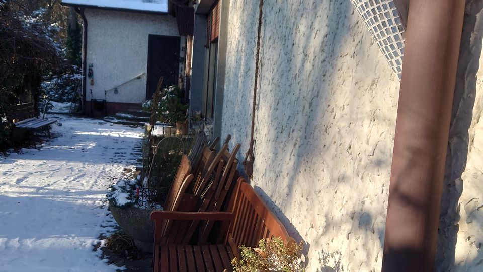 Ein kleiner, schneebedeckter Gartenbereich mit einer Hauswand aus weiß verputztem Stein, einer Reihe Holzstühle und einem runden Tisch, auf dem rot-gelbe Äpfel, ein Pflanztopf und eine Schale mit Kiefernzapfen stehen. Im Hintergrund liegt ein Nebengebäude mit einem schneeüberzogenen Dach, umgeben von hohen Bäumen.