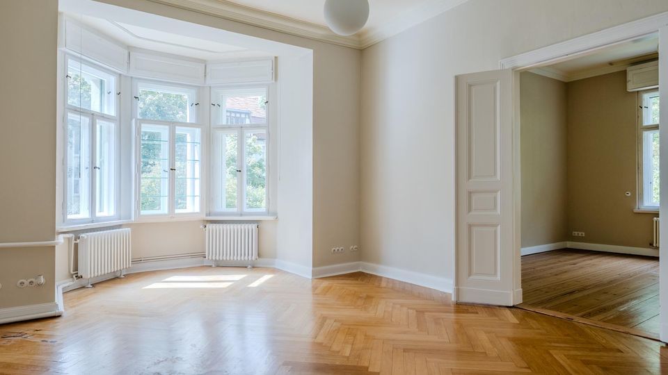 Heller, leerer Altbauwohnraum mit Fischgrätparkett, hohen Decken und großem Erker mit weißen Sprossenfenstern und Heizkörpern darunter. Rechts führt eine offene Kassettentür in ein angrenzendes Zimmer mit Holzboden und weiterem Fenster.
