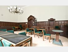 Innenraum eines historischen Gerichtssaals mit holzvertäfelten Wänden, schlichten Holztischen und grünen Polsterstühlen. Im Hintergrund befindet sich eine dekorative Holztür mit geschwungenem Türrahmen und eine erhöhte Richterbank.