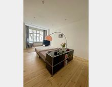 Helles Schlafzimmer mit Holzfußboden, einem Bett in der Mitte, schwarzem Regalschrank im Vordergrund und einer großen, gebogenen Stehlampe mit orangefarbener Schirm. Im Hintergrund steht ein Schreibtisch vor einem Fenster mit grauen Vorhängen.