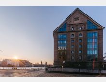 Backsteingebäude mit der Aufschrift „Osthafen Speicher“, kombiniert mit modernen Glasfassaden, am Ufer eines Flusses. Im Hintergrund sind weitere Gebäude, eine Brücke und zwei Türme zu sehen, während die tiefstehende Sonne die Szene beleuchtet.