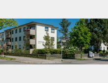 Wohngebäude in einer ruhigen Wohnstraße, bestehend aus einem weiß verputzten, zweigeschossigen Haus mit Balkonen, teilweise mit Blumenkästen verziert. Umgeben von Bäumen, Sträuchern und einem Holzzaun, mit einem Straßenschild und einem Bürgersteig im Vordergrund.