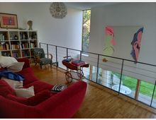 Wohnzimmer auf einer Galerieebene mit moderner Einrichtung, darunter ein rotes Sofa, ein Bücherregal und ein blumig gemusterter Sessel. Eine große Fensterfront bietet Blick auf einen Garten und ein buntes Kunstwerk hängt an der weißen Wand.