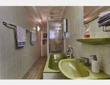 Badezimmer mit pastellfarbenen Kacheln an den Wänden, ausgestattet mit einer grünen Badewanne, einem grünen Waschbecken und einer Duschkabine mit Glaswänden. Im Hintergrund befindet sich ein kleines Regal und ein Fenster mit milchigem Glas.