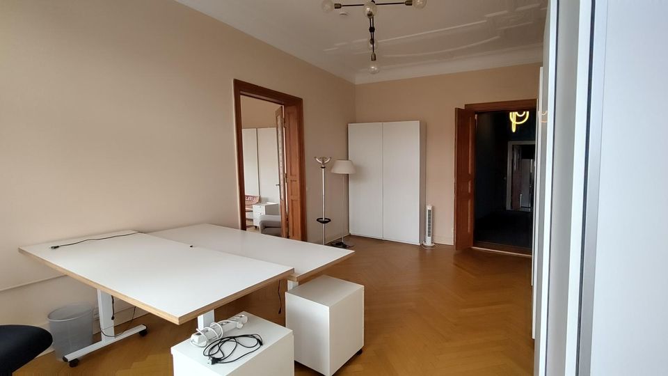 Heller Büroraum mit hellem Holzboden, großen weißen Schreibtischen und schlichten weißen Schränken sowie Deckenleuchte. Zwei Holztüren führen in angrenzende Räume, an der rechten Seite ist ein Fenster- oder Glaselement mit Rahmen zu sehen.