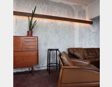 Industrial anmutender Wohnraum mit rauer Betonwand und einer langen Holzleiste mit indirekter Beleuchtung. Im Vordergrund stehen ein braunes Ledersofa und ein Ledersessel, daneben ein schwarzer Barhocker und ein Holzsideboard mit Topfpflanze.