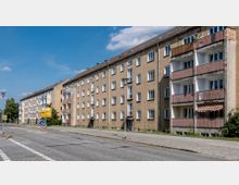 Eine Reihe mehrstöckiger Wohngebäude im schlichten Stil, mit verputzter Fassade und Balkonen, die teils renovierungsbedürftig aussehen. Im Vordergrund eine ruhige Straße mit Gehwegen, Straßenschildern und vereinzelten Laternen, umgeben von einem klaren blauen Himmel.
