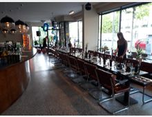 Innenraum eines modernen Restaurants mit großen Fenstern, die Blick auf die Straße bieten. Links befindet sich eine geschwungene Theke, rechts stehen längliche, gedeckte Tische mit Gläsern, Geschirr und Dekorationen, umgeben von braunen Stühlen.