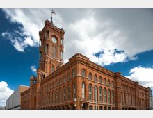 Backsteingebäude mit markantem Turm und Uhr, bekannt als das Rote Rathaus. Die Fassade ist mit Rundbogenfenstern gestaltet, und eine Fahne weht auf der Spitze des Turms; der Himmel ist teilweise bewölkt.