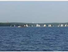 Ein großer See mit einer Vielzahl von Segelbooten, umgeben von bewaldeten Ufern. Im Hintergrund sind ein Turm und weitere entfernte Gebäude sichtbar.