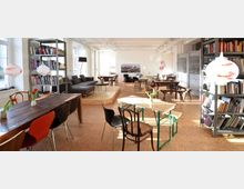 Heller Raum mit skandinavisch-modernem Interieur, bestehend aus Holz- und Metalltischen, gemischten Sitzgelegenheiten und einer Sofaecke auf einem Podest. An den Wänden befinden sich Bücherregale, dekorative Lampen hängen von der Decke, und durch große Fenster fällt viel Tageslicht.