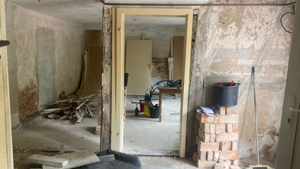 Ein renovierungsbedürftiger Innenraum mit freiliegenden Backsteinwänden, abblätterndem Putz und Werkzeugen. Im Vordergrund liegen Baumaterialien und ein Eimer steht auf einem Stapel Ziegelsteine.