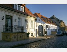 Eine gepflasterte Straße mit Reihenhäusern im historischen Baustil, gekennzeichnet durch helle Fassaden, rote Ziegeldächer und dekorative Balkongeländer. Einige Passanten und parkende Fahrzeuge sind am Straßenrand zu sehen, während Laternenmasten die Straße säumen.