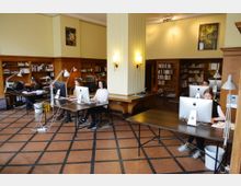 Ein heller Büroraum mit hohen Decken, in dem mehrere Personen an Schreibtischen mit großen Desktop-Computern arbeiten. Holzregale mit Büchern und Akten umrahmen den Raum, der mit gemustertem Steinboden und Wandlampen gestaltet ist.