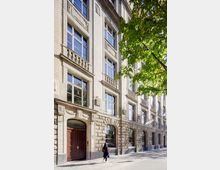 Mehrstöckiges, historisches Stadthaus mit heller Backsteinfassade und rustiziertem Sockelgeschoss, großen rechteckigen Fenstern und einem Rundbogen-Eingang. Im Vordergrund verläuft ein Gehweg entlang der Straße, rechts ragen grüne Baumkronen ins Bild.