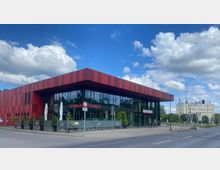 Modernes Gebäude mit rot-schwarz gestreifter Fassade und großen Glasfronten, das als „Bürgerhaus“ gekennzeichnet ist. Vor dem Gebäude befinden sich eine Terrasse mit Sonnenschirmen und Pflanzen sowie ein Gehweg, im Hintergrund ist eine Straße mit weiteren Gebäuden sichtbar.
