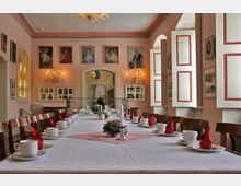 Langer, festlich gedeckter Tisch in einem historischen Raum mit rosa Wänden. Der Raum ist geschmückt mit Gemälden, Kronleuchtern und dekorativen Fenstern mit roten Rahmen; im Hintergrund ist eine offene Tür zu sehen.