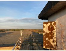 Flaches Dach eines Gebäudes mit Betonoberfläche und Geländer, umgeben von herbstlichem Wald. An der rechten Seite steht eine beschädigte Tür mit rostigen und abblätternden Farben, während sich im Hintergrund ein weiter Himmel zeigt.