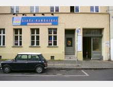 Fassade eines gelb verputzten Gebäudes mit einem blauen Schild mit der Aufschrift „Café Große Hamburger - Ein Projekt der Caritas-Suchtberatung“. Vor dem Haus parkt ein schwarzer Kleinwagen, daneben befindet sich eine Eingangstür und eine Durchfahrt mit Tor.