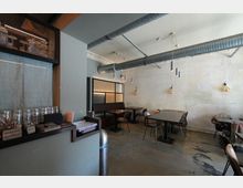 Innenraum eines industriell gestalteten Cafés oder Restaurants mit hohen Decken, sichtbaren Lüftungsrohren und hängenden Leuchten. Links eine Theke mit gestapelten Gläsern, rechts mehrere einfache Tische und Stühle vor einer hellen, roh verputzten Wand.