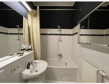 Weiß gefliestes Badezimmer mit schwarzer Decke, einer Badewanne mit Duscharmatur, einem cremefarbenen Duschvorhang, einem Waschbecken mit Spiegel sowie einem Heizkörper an der Wand. Auf der Ablage befinden sich Zahnbürsten, Haarbürsten und Pflegeprodukte.