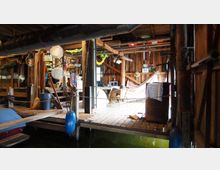 Holzinterieur eines rustikalen Bootshauses, mit hölzernem Steg über Wasser, von Ketten und Fendern gesichert. Im Innenraum befinden sich Hängematten, Stühle, dekorative Elemente und verschiedene Gegenstände wie Eimer, Besen und Laternen.