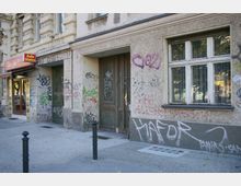 Fassade eines Altbaus mit grauem Putz, besprüht mit zahlreichen Graffiti. Links befindet sich ein Eingang zu einem kleinen Asia-Bistro mit Werbeschild und Markise, daneben eine große Holztür und ein Fenster mit Gardinen.