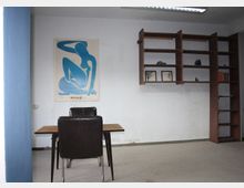 Ein schlicht eingerichteter Raum mit hellem Teppichboden, einem Holztisch und zwei dunklen Stühlen im Vordergrund. An der Wand hängt ein Kunstplakat mit einer blauen Silhouette und daneben ein Regal aus dunklem Holz mit Büchern, kleinen Dekorationsgegenständen und zwei gerahmten Bildern.