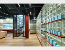 Innenraum einer modernen Apotheke mit Produkten auf roten Regalen vor einer Wand aus Glasbausteinen. Der Raum ist hell beleuchtet, hat eine Holzoptik am Boden und verfügt über ein gläsernes Schrankelement in der Mitte.