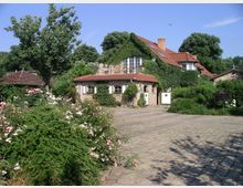 Ein ländliches Haus aus Naturstein mit roten Ziegeldächern, teilweise von Efeu bewachsen, umgeben von einem gepflegten Garten mit Blumen, Bäumen und Sträuchern. Im Vordergrund ist ein gepflasterter Hof zu sehen, der das Gebäude umgibt.