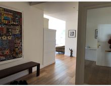 Flur in einer modernen Wohnung mit Holzboden und weiß gestrichenen Wänden. An der linken Wand hängt ein großes, farbenfrohes Kunstwerk, gegenüber steht eine dunkle Holzbank. Im Hintergrund sind eine helle Sitzecke mit Blick nach draußen und eine geöffnete Zimmertür sichtbar.