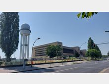 Straßenansicht mit einem großen, rechteckigen Gebäude im Hintergrund, das eine schlichte Fassade und Reihen gleichförmiger Fenster aufweist. Links daneben steht ein hoher, weißer Turm mit einer kugelförmigen Struktur an der Spitze, umgeben von Bäumen und einem umzäunten Grünbereich.