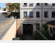 Eingang zu einem Hofdurchgang mit Schild „Kurt Mühlenhaupt Museum“ an einem weiß gestrichenen Backsteingebäude mit Rundbogenfenstern und teils herabgelassenen Rollläden. Links ist eine flache Dachfläche mit Kies und Pflanzenkübeln zu sehen, rechts begrenzen Bewuchs und eine Lichterkette den schmalen Zugang.