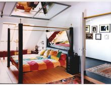 Schlafzimmer mit einem bunten Himmelbett und Holzboden, darüber ein Spiegel an der Decke. Im Hintergrund befindet sich ein Flur mit Kunstwerken an weißen Wänden.