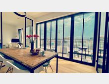 Heller, moderner Wohnraum mit einem großen Holztisch, weißen Stühlen und einer Arbeitsstation im Hintergrund. Durch bodentiefe Fenster ist eine Terrasse mit Sitzgelegenheiten zu sehen, im Hintergrund ragt der Berliner Fernsehturm in den Himmel.