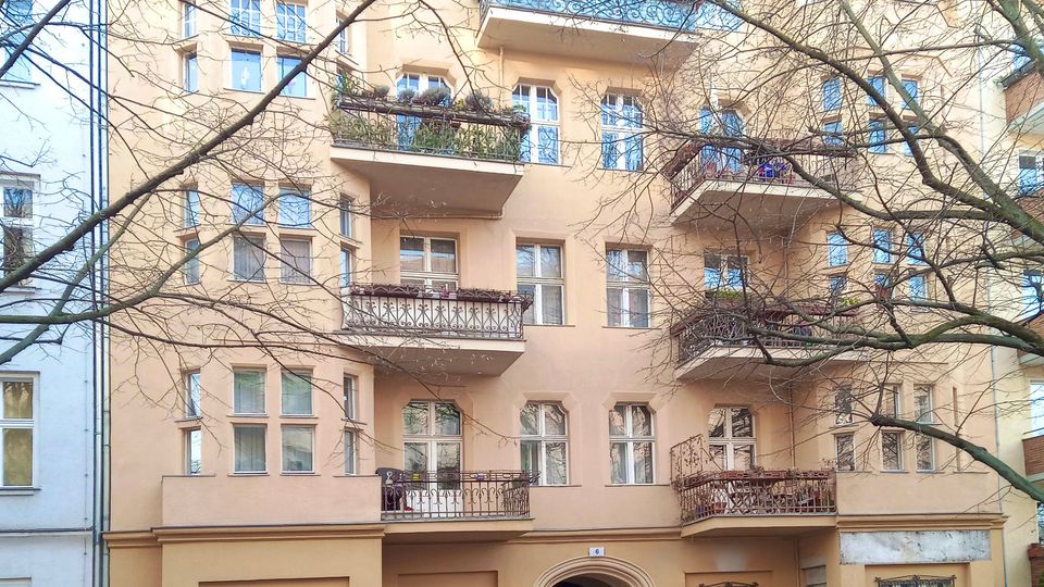 Mehrstöckiges Wohnhaus mit hellbeiger Fassade, roten Ziegeldach und mehreren Balkonen mit schmiedeeisernen Geländern. Im Vordergrund stehen geparkte Autos an einer Kopfsteinpflasterstraße, teilweise verdeckt von kahlen Baumästen.
