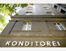 Fassade eines mehrstöckigen Gebäudes aus dunkelgrauem Putz mit weiß gerahmten Fenstern und dem Schriftzug „Konditorei“ in großen weißen Buchstaben im unteren Bereich. Im Vordergrund sind grüne Baumzweige sichtbar.