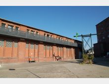 Backstein-Gebäude mit großen Glasbausteinfenstern und einer langen Metallbrücke im oberen Bereich. In der rechten Bildhälfte befindet sich eine grüne Statue auf einem Metallträger, der in den Hof ragt.