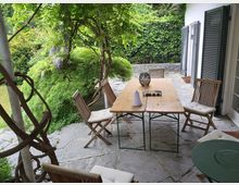 Steinterrasse eines Gartens mit einem Holztisch, umgeben von Holzstühlen mit Sitzpolstern. Im Hintergrund sind üppige grüne Pflanzen, ein Baum sowie eine mit Efeu bedeckte Steinmauer zu sehen; links rankt eine Pflanze an einer Pergola, rechts schließen sich ein Gebäudeteil mit schwarzen Fensterläden und eine Tür an.