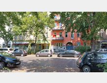 Straßenszene mit Wohn- und Geschäftshäusern, darunter ein orangefarbenes Gebäude mit Graffiti und Balkonen. Vor den Häusern stehen parkende Autos, und schattenspendende Bäume säumen die gepflasterte Straße.