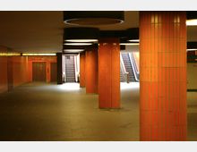 Unterirdischer U-Bahn-Bereich mit orangefarbenen, gefliesten Säulen, schwarzen runden Deckenleuchten und Rolltreppen im Hintergrund. Links ist eine Tür zu sehen, die wie ein Aufzug wirkt, zudem gibt es ein leuchtendes Schild mit der Aufschrift „Aufzug“.