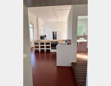 Ein moderner, heller Raum mit offener Küche, weiß gestrichenen Wänden und rotem Boden. Links befinden sich große gewölbte Fenster, und im Vordergrund ist eine Küchenzeile mit offenen Regalfächern und einem Herd zu sehen; daneben führt eine Tür zu einem Badezimmer mit roten Mosaikfliesen.