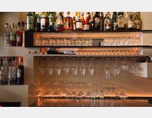 Regal mit verschiedenen Flaschen Alkohol oben, darunter mehrere Reihen aufgehängter Weingläser und aufgestellte Trinkgläser; moderne Bar-Einrichtung mit warmer Beleuchtung.