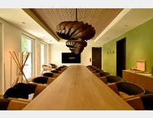 Moderner Konferenzraum mit langem Holztisch und vielen Stühlen, großen Fenstern an der linken Seite und einem Wandbildschirm am Ende des Raums. Auffällig sind die skulpturale, dunkle Pendelleuchte und die Holzlamellendecke; rechts eine grüne Akzentwand mit Türen und Sideboard.